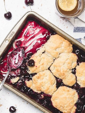 A baking dish of cherry cobbler made with an easy cherry pie casserole recipe sits on a white surface, with servings on plates, fresh cherries, and a spoon nearby.