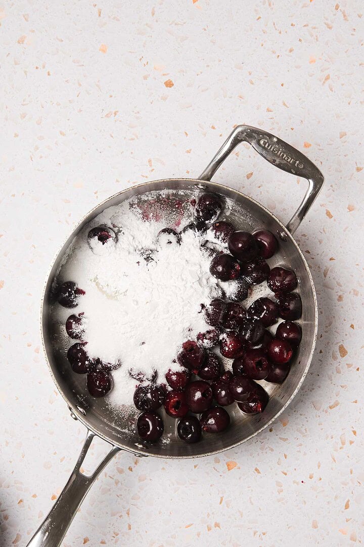 A saucepan with fresh cherries and a mound of white powdered sugar hints at an easy recipe for Cherry Pie on a light speckled surface.