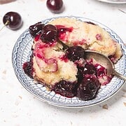 A plate of cherry cobbler with a spoon, fresh cherries, and another plate in the background—a delicious cherry dessert that's perfect for any occasion.