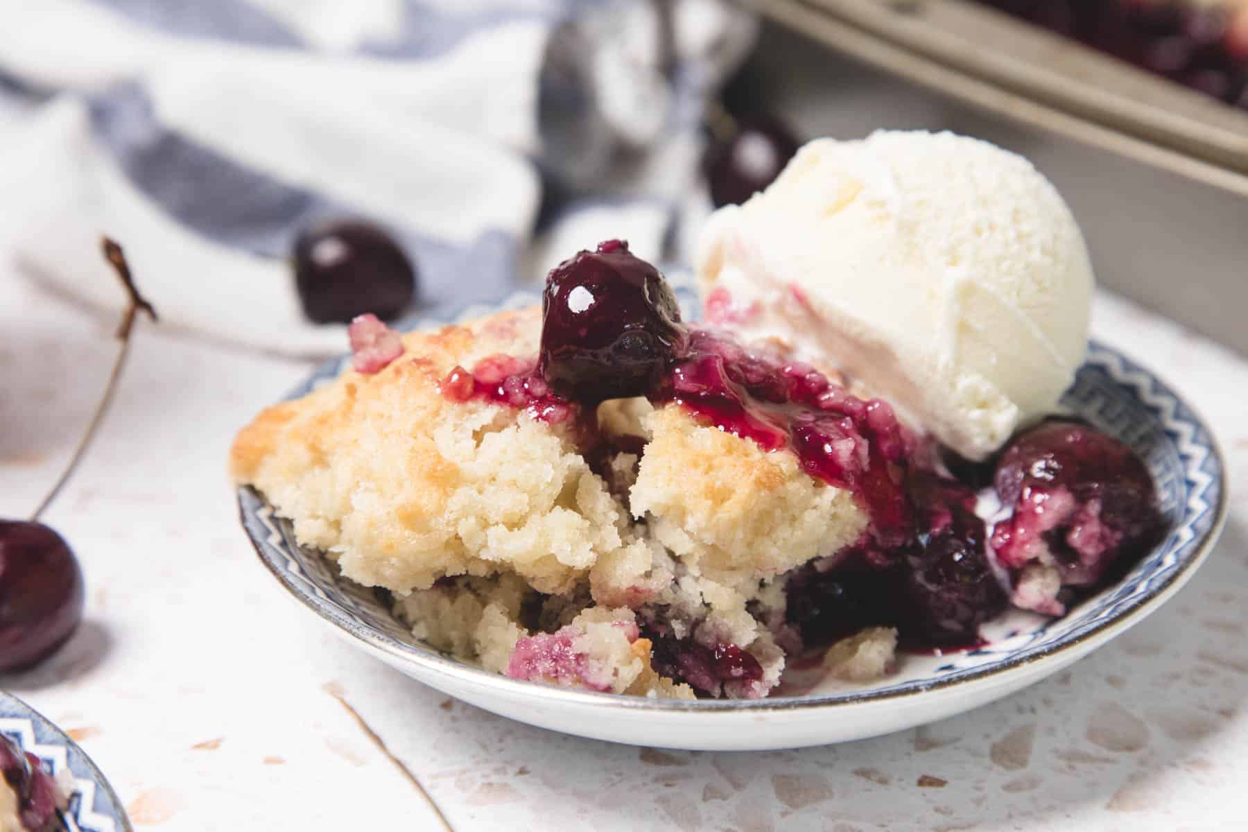A plate of cherry cobbler with a scoop of vanilla ice cream, garnished with fresh cherries&mdash;an easy recipe inspired by classic cherry pie.