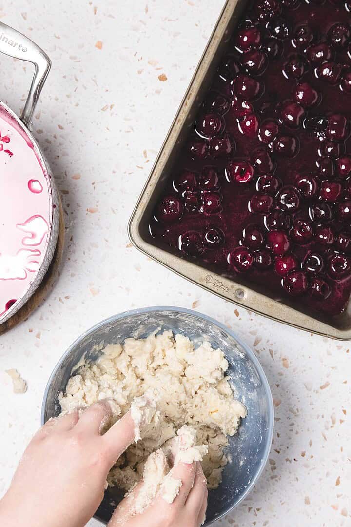 Hands mixing dough in a bowl next to a pan of cherry filling on a kitchen counter, preparing an easy cherry pie recipe.