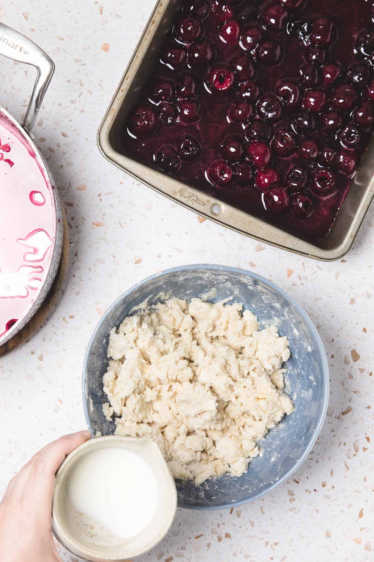 A bowl of dough, a pan of cherry filling, and a hand holding a small pitcher of milk on the counter&mdash;everything you need for an easy Cherry Pie Casserole recipe.