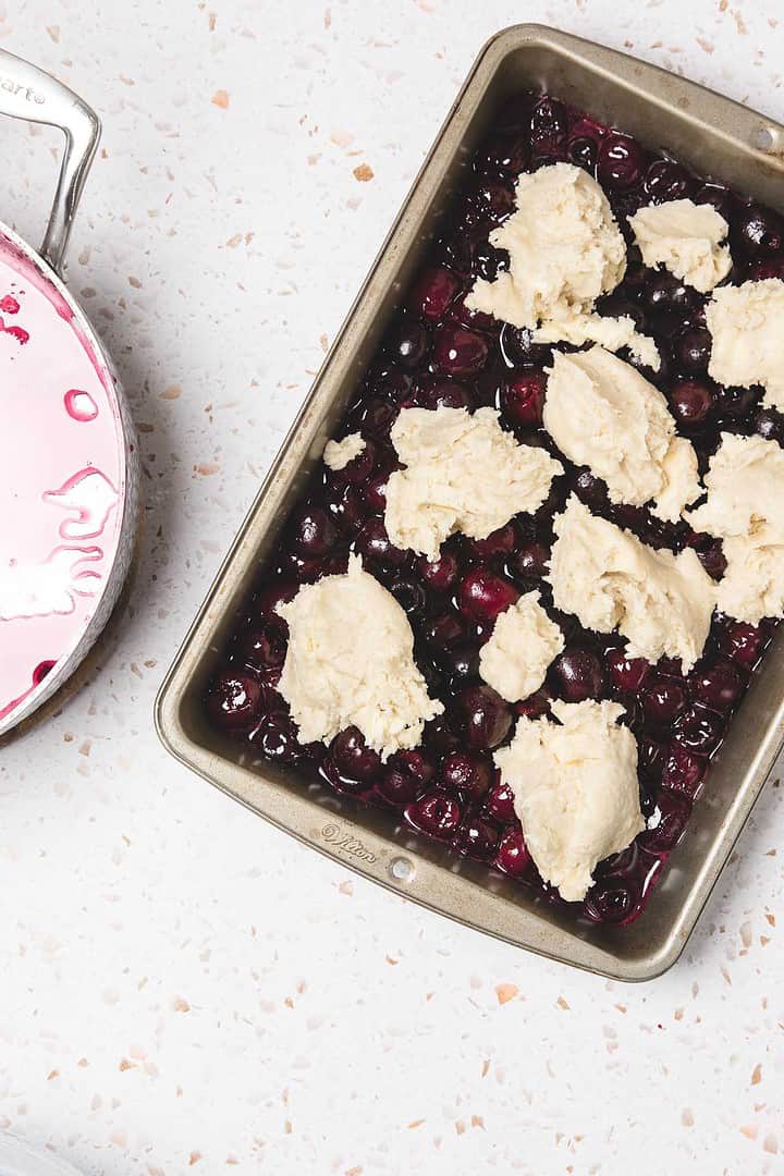 A baking pan with cherry pie casserole&mdash;layers of cherry filling and biscuit dough&mdash;sits beside a pot of cherry sauce on a white counter, showcasing an easy cherry pie recipe.