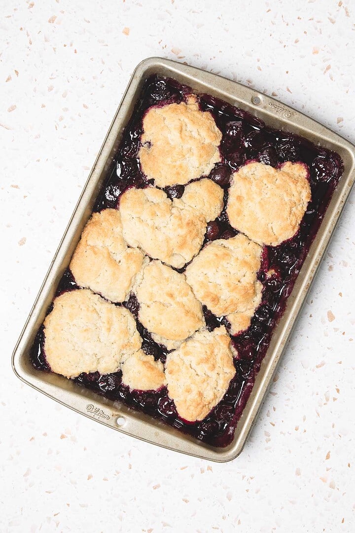 A baking pan filled with blueberry cobbler, reminiscent of an Easy Cherry Pie, topped with golden, flaky biscuits on a light countertop.