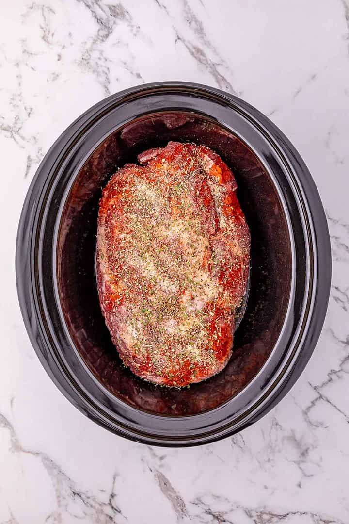 Seasoned raw meat in a black slow cooker on a white marble countertop, ready to be cooked for an Easy Pork Roast with potatoes and carrots.