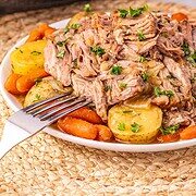 Plate of shredded Crock Pot Pork Roast with potatoes and carrots, garnished with parsley, in front of a slow cooker.