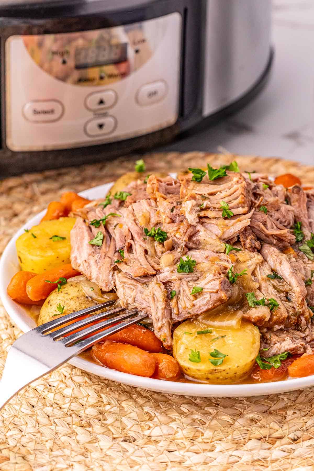 Plate of tender Crock Pot Pork Roast, shredded with potatoes and carrots, garnished with parsley, in front of a slow cooker.