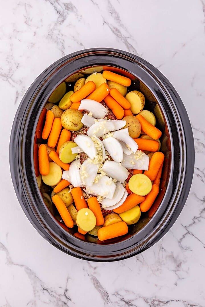 Sliced onions, baby carrots, potatoes, and minced garlic in a Crock Pot Pork Roast setting on a marble countertop.