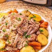 A plate of shredded Crock Pot Pork Roast with potatoes and carrots, garnished with parsley, sits in front of a slow cooker.