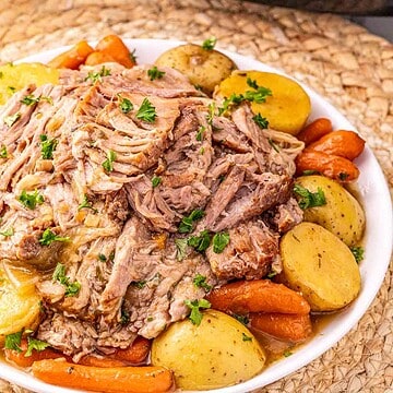 A plate of shredded Crock Pot Pork Roast with potatoes and carrots, garnished with parsley, sits in front of a slow cooker.