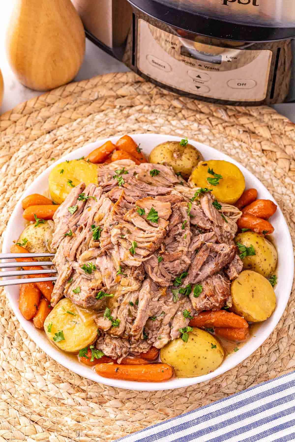 Shredded Crock Pot Pork Roast with potatoes and carrots, garnished with parsley, served on a white plate beside a slow cooker for an easy pork roast dinner.