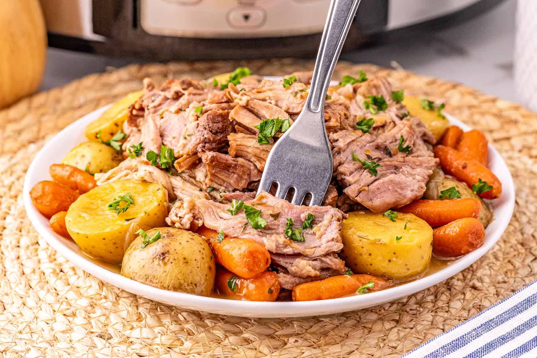 Plate of Crock Pot Pork Roast with carrots, potatoes, and herbs, a fork in the tender meat; slow cooker in background.