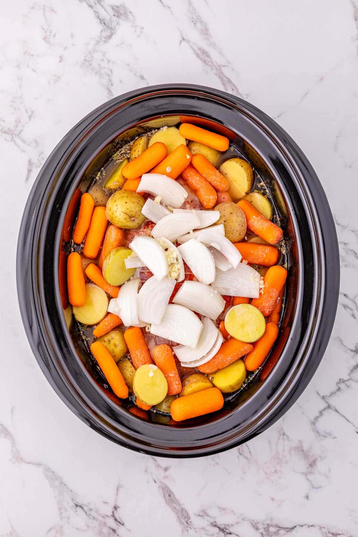 Crock Pot Pork Roast with potatoes and carrots, plus onions and garlic, ready to cook in a slow cooker on a marble surface.