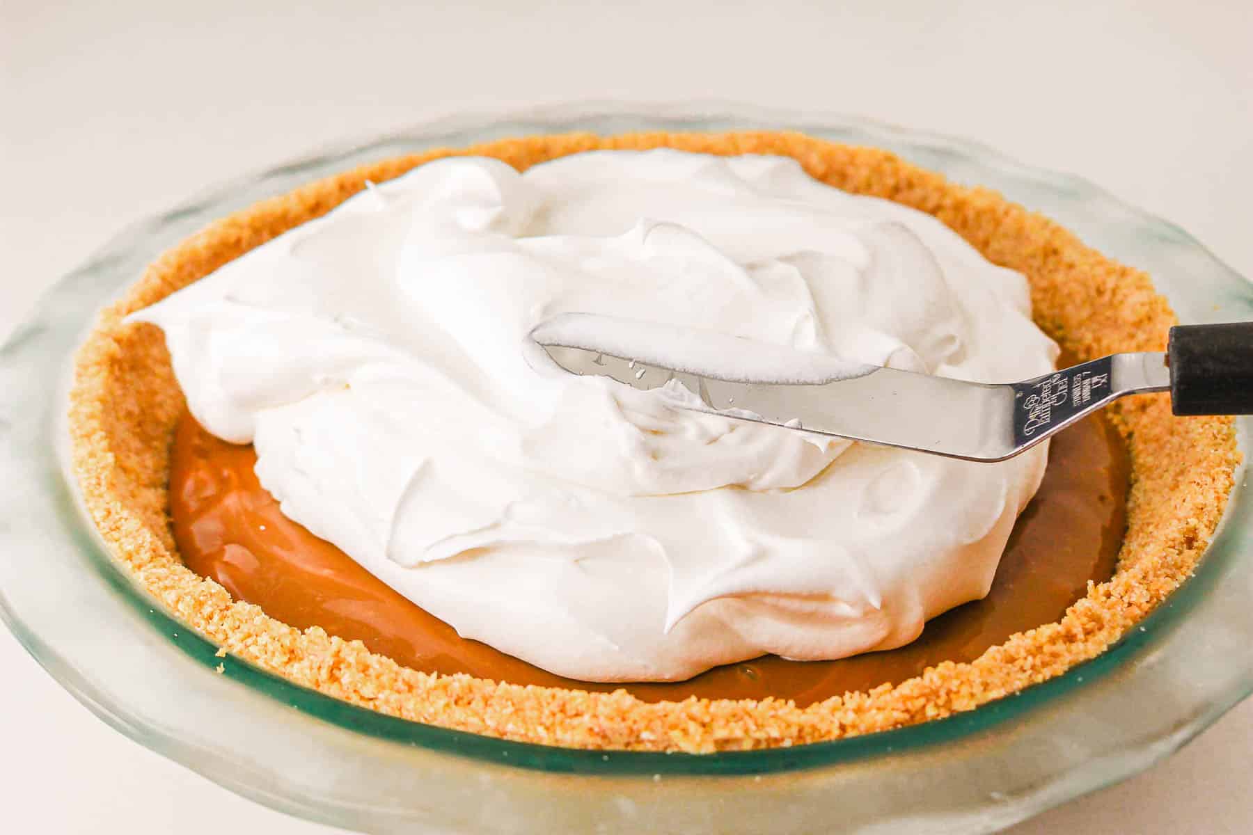A caramel pie with a graham cracker crust is being topped with whipped cream using a spatula—an irresistible recipe you'll want to make again and again.