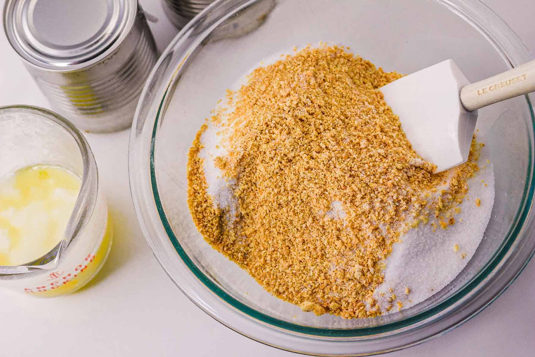A bowl of graham cracker crumbs and sugar with a spatula, next to melted butter and canned ingredients—perfect for starting a Slow Cooker Caramel Pie recipe.