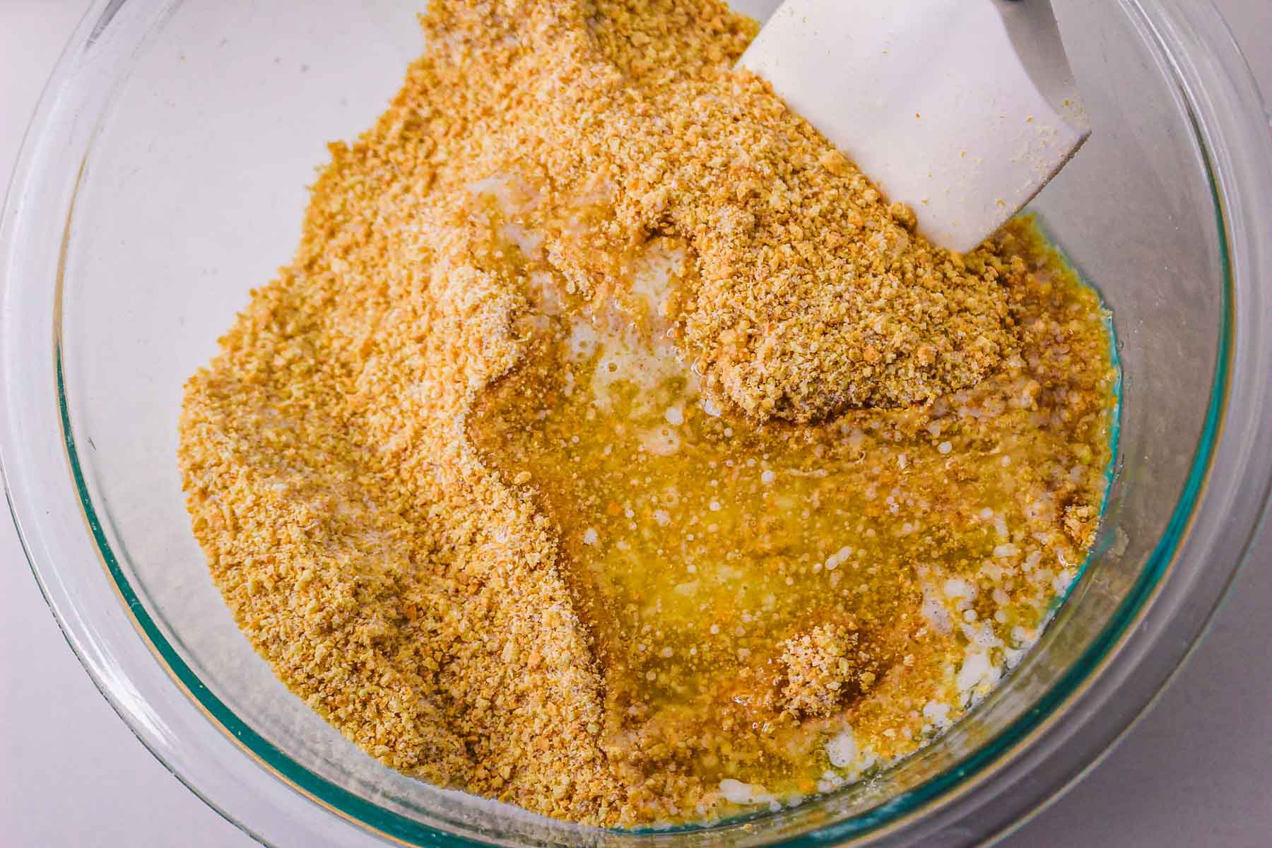 Crushed graham crackers mixed with melted butter and sugar in a glass bowl with a white spatula, forming the perfect base for a delicious caramel pie recipe.