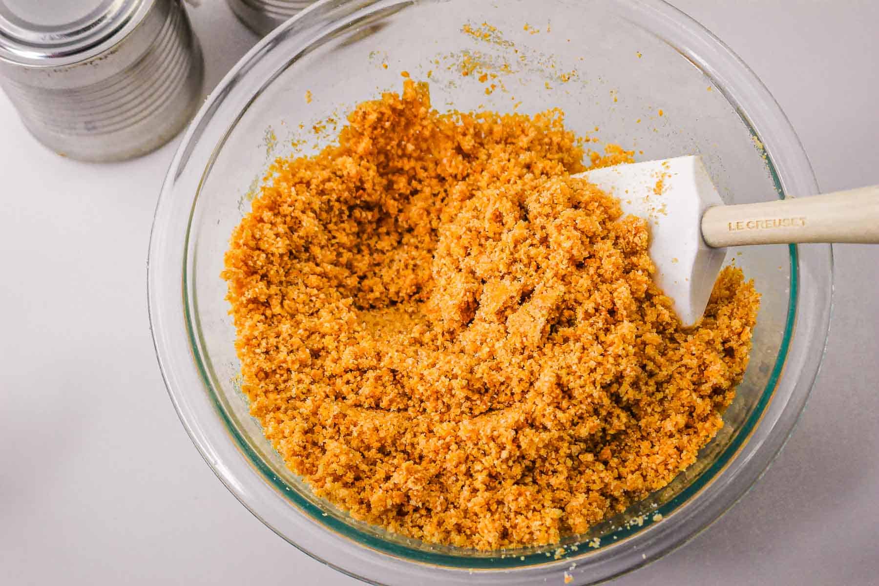 Crushed graham crackers mixed with butter in a glass bowl, stirred with a white spatula—perfect for a Caramel Pie recipe.