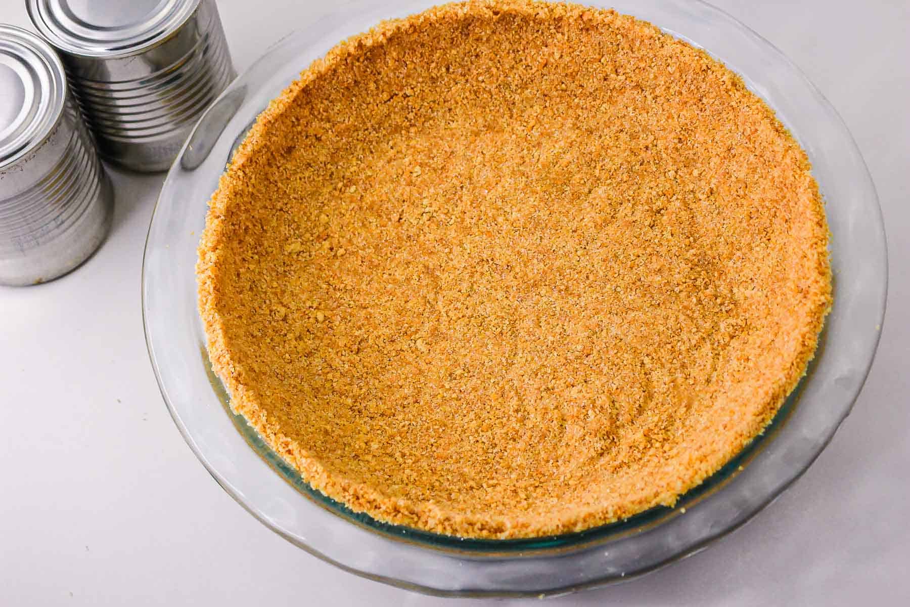 A glass pie dish with a pressed graham cracker crust, perfect for a caramel pie recipe, sits next to a few tin cans on a white surface.