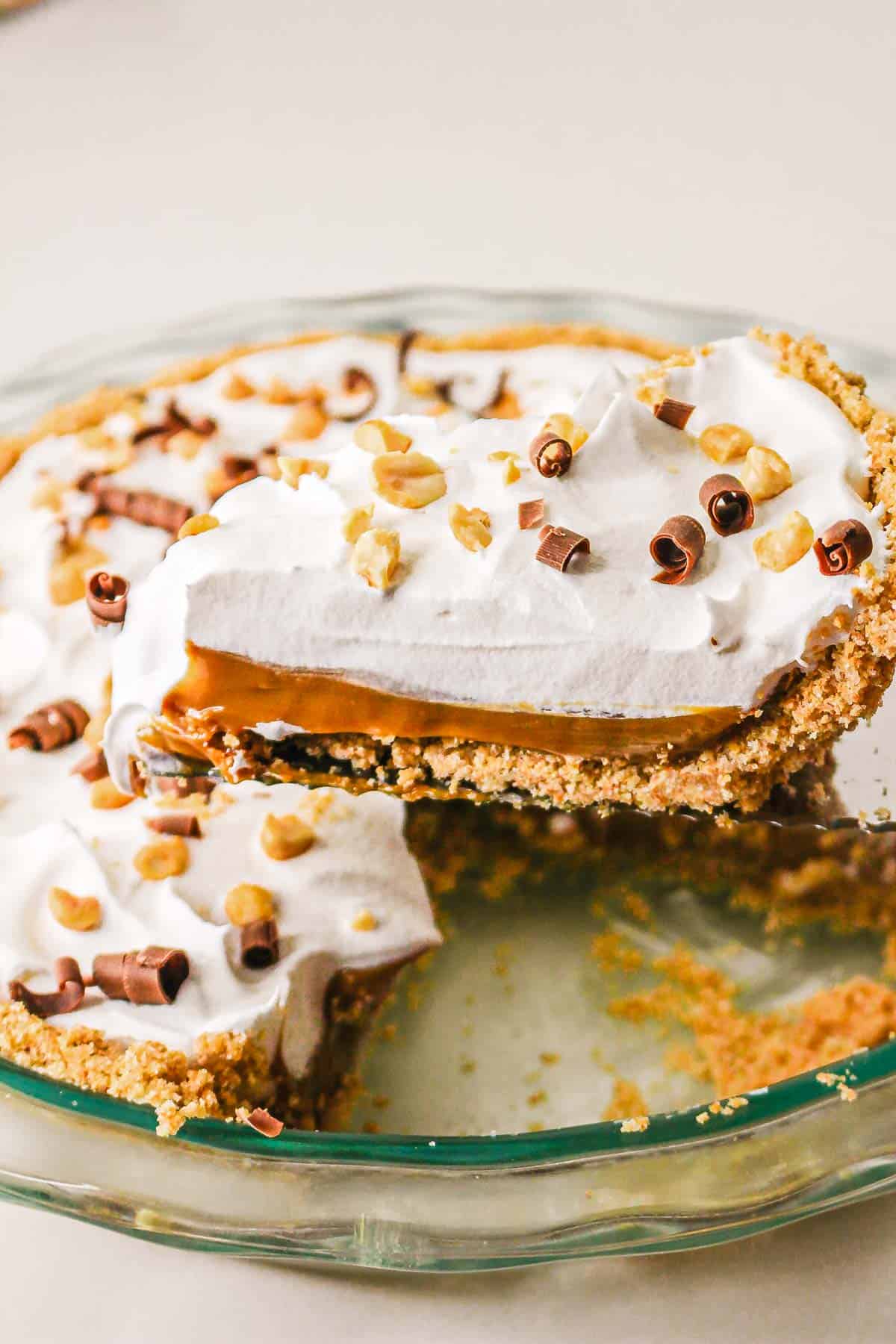 A Caramel Pie with a graham crust, caramel filling, whipped cream, chocolate curls, and chopped nuts sits with a slice missing—an irresistible slow cooker recipe for dessert lovers.