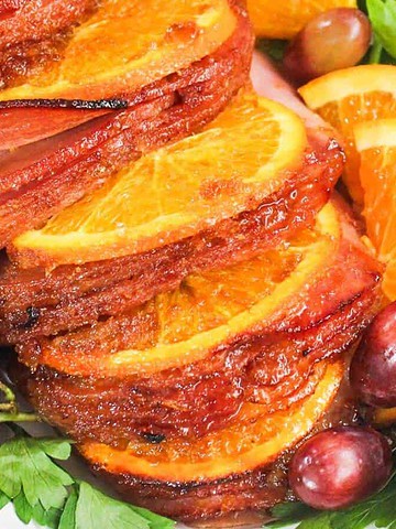 Sliced glazed ham garnished with orange slices, red grapes, and fresh parsley on a platter.