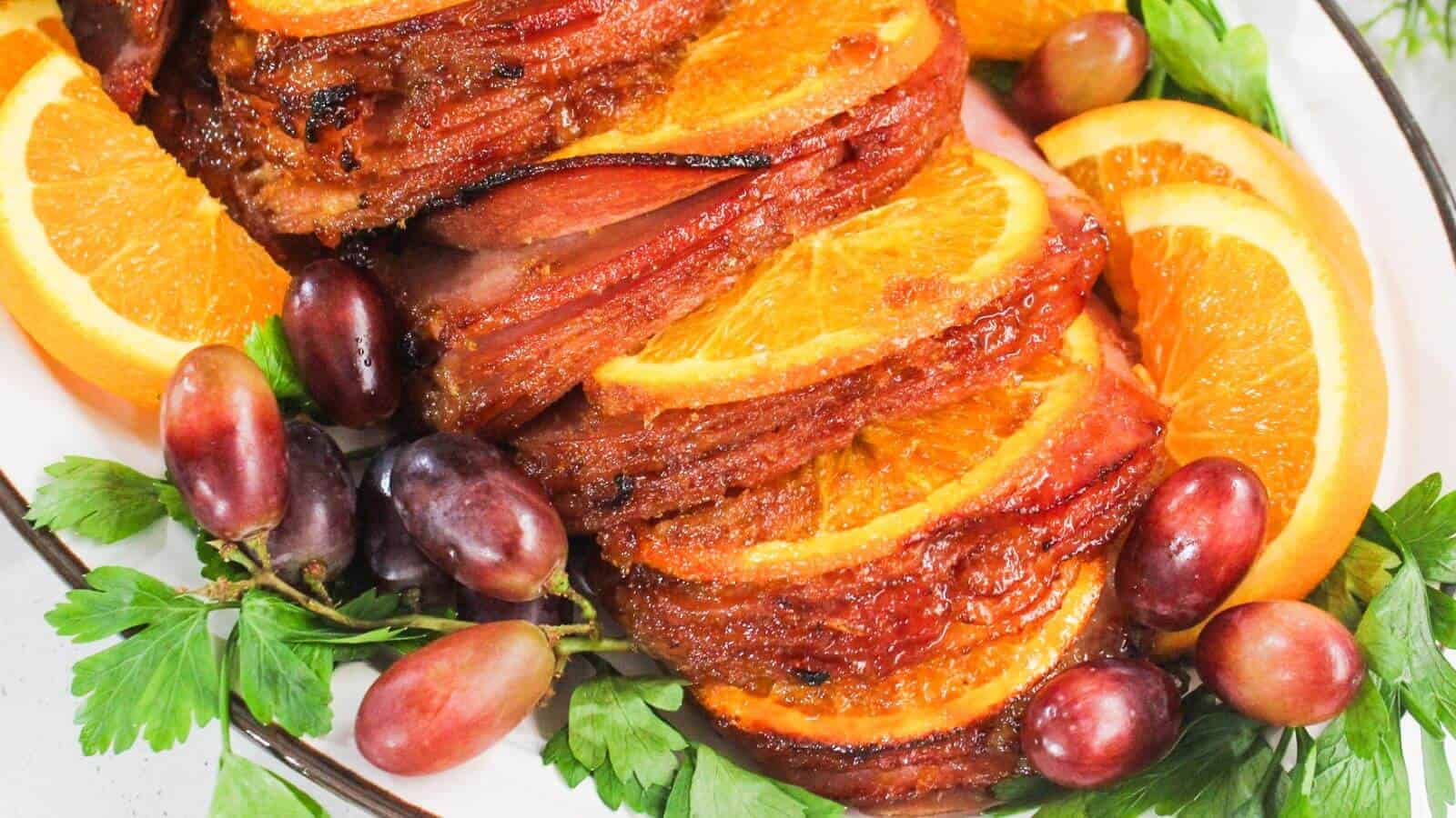 Sliced glazed ham garnished with orange slices, red grapes, and fresh parsley on a platter.