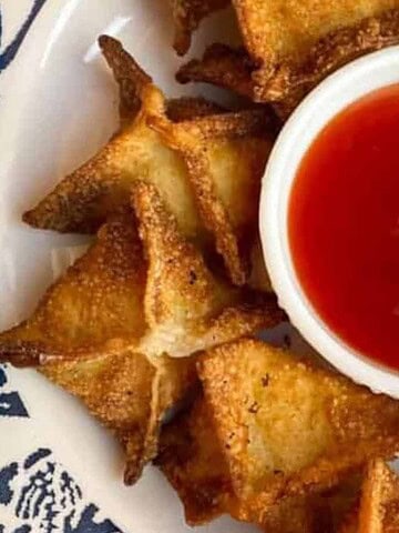 Golden fried wontons arranged in a circle around a bowl of red dipping sauce on a patterned plate.