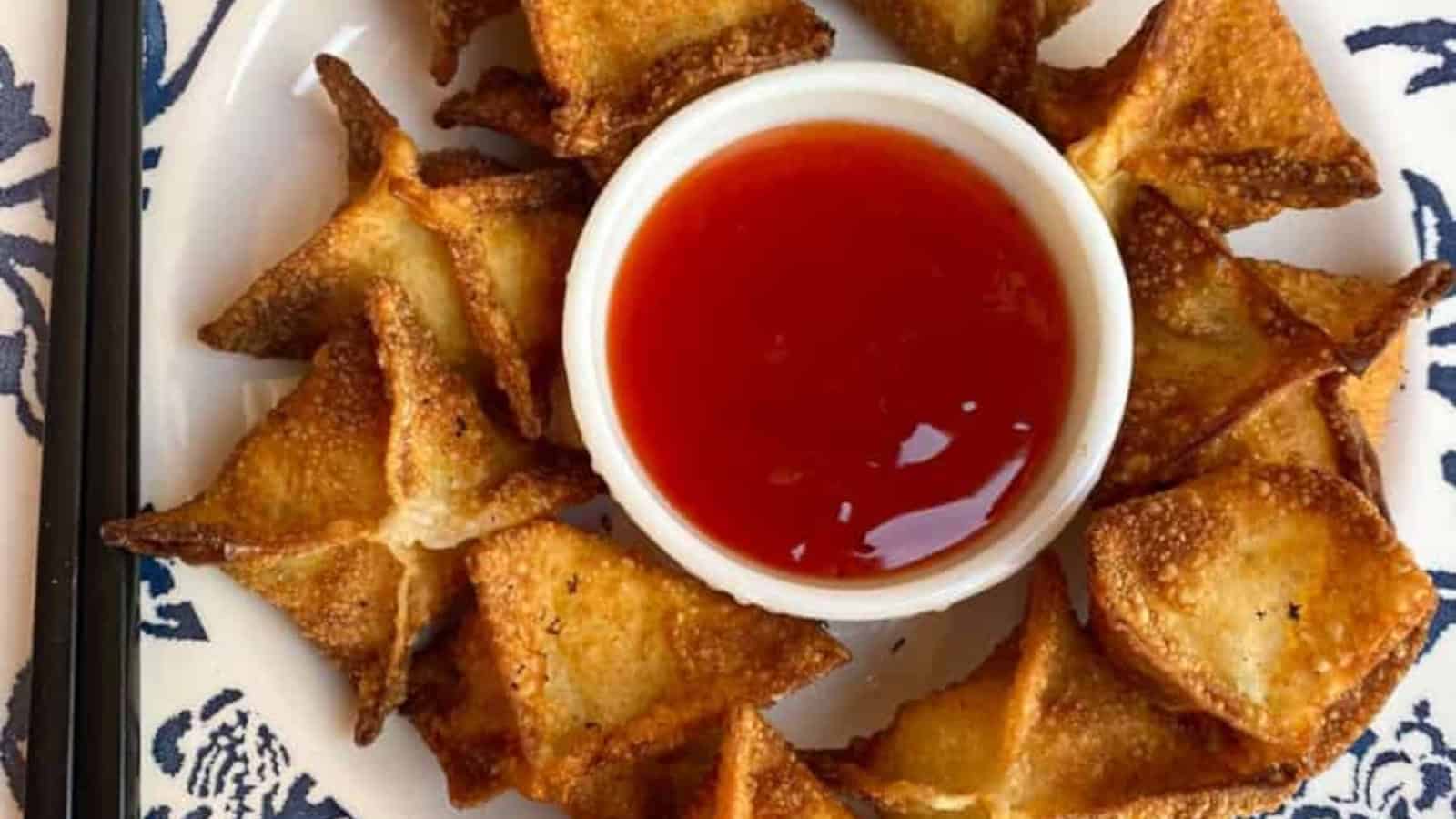 Golden fried wontons arranged in a circle around a bowl of red dipping sauce on a patterned plate.