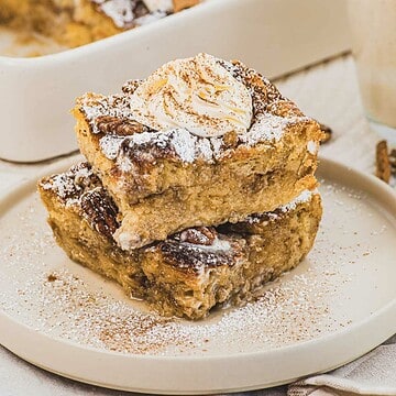 Two pieces of Eggnog French Toast casserole stacked on a plate, topped with whipped cream and sprinkled with powdered sugar.