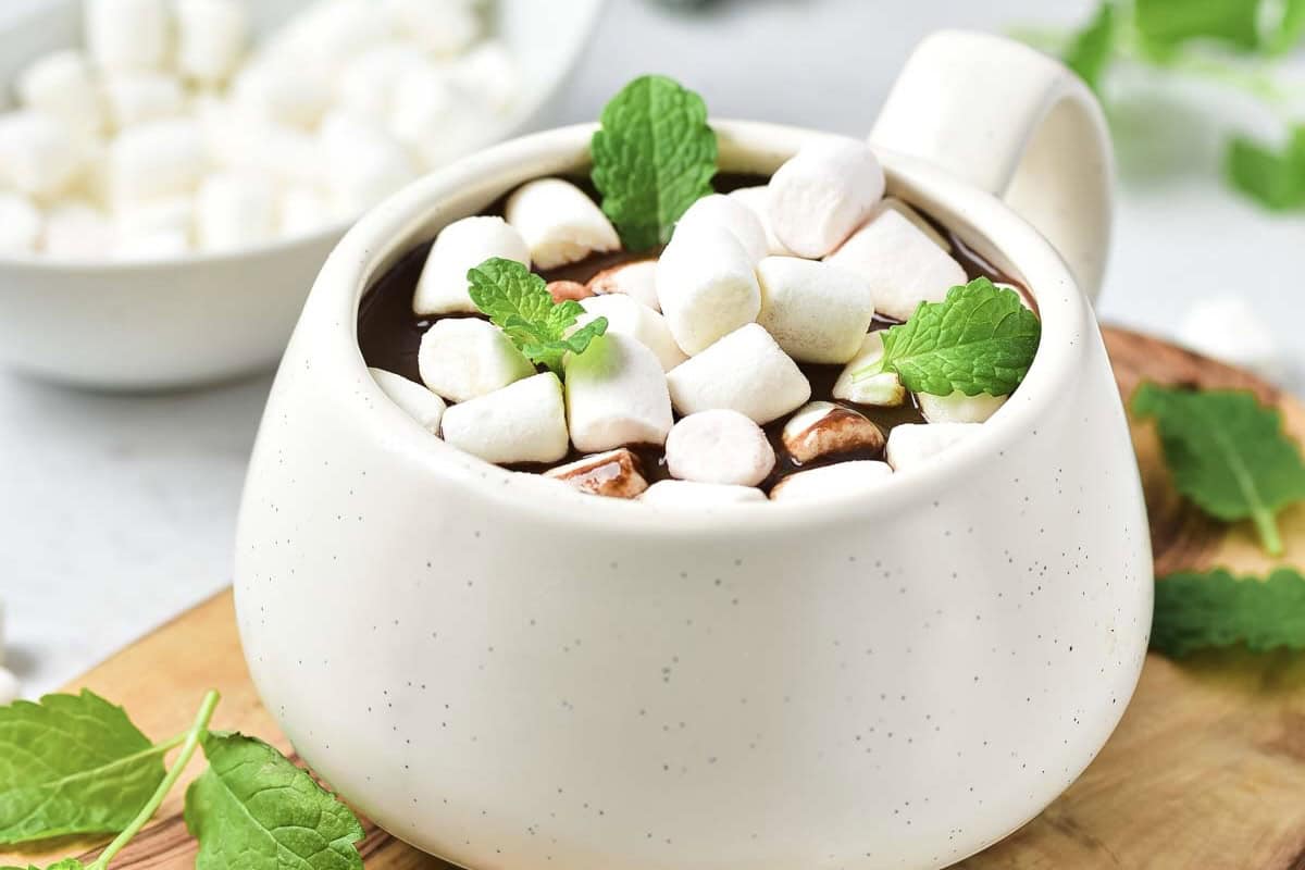 A mug of rich peppermint hot chocolate topped with mini marshmallows and mint leaves sits invitingly on a wooden board.
