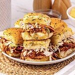 Pulled BBQ chicken and coleslaw sliders stacked on a white plate with golden, seasoned buns.