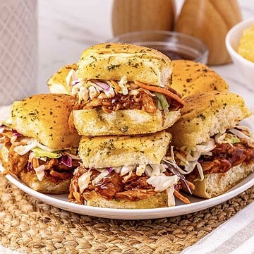 Pulled BBQ chicken and coleslaw sliders stacked on a white plate with golden, seasoned buns.