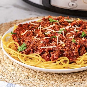 A plate of Spaghetti Bolognese topped with meat sauce, grated cheese, and parsley sits on a woven placemat.