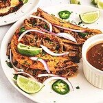 Three slow cooker birria tacos with shredded meat, lime wedges, sliced onions, jalapeños, and dipping sauce on a plate.