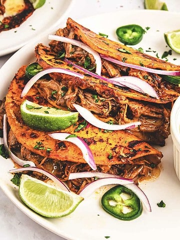 Three slow cooker birria tacos with shredded meat, lime wedges, sliced onions, jalapeños, and dipping sauce on a plate.