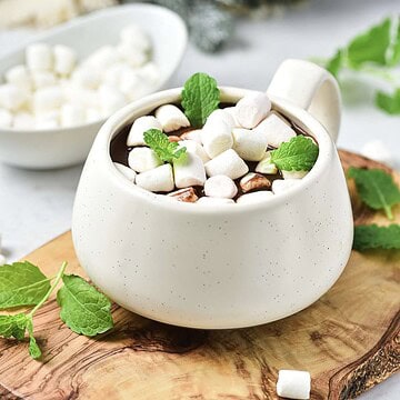 A mug of hot chocolate topped with marshmallows and mint leaves on a wooden board.