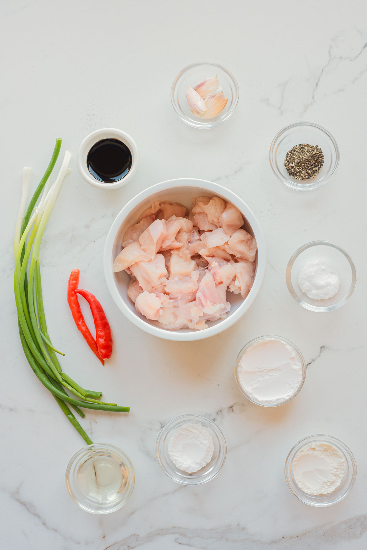 Raw chicken pieces, green onions, red chili, and seasonings in small bowls arranged on a white marble surface, ready to make delicious Air Fryer Chicken.