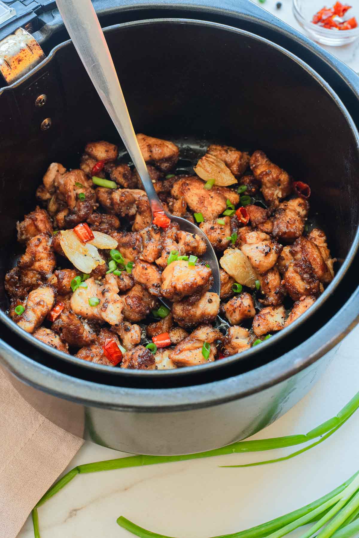 Salt and pepper chicken pieces with vegetables in an air fryer basket, perfectly cooked and seasoned, with a serving spoon inside.