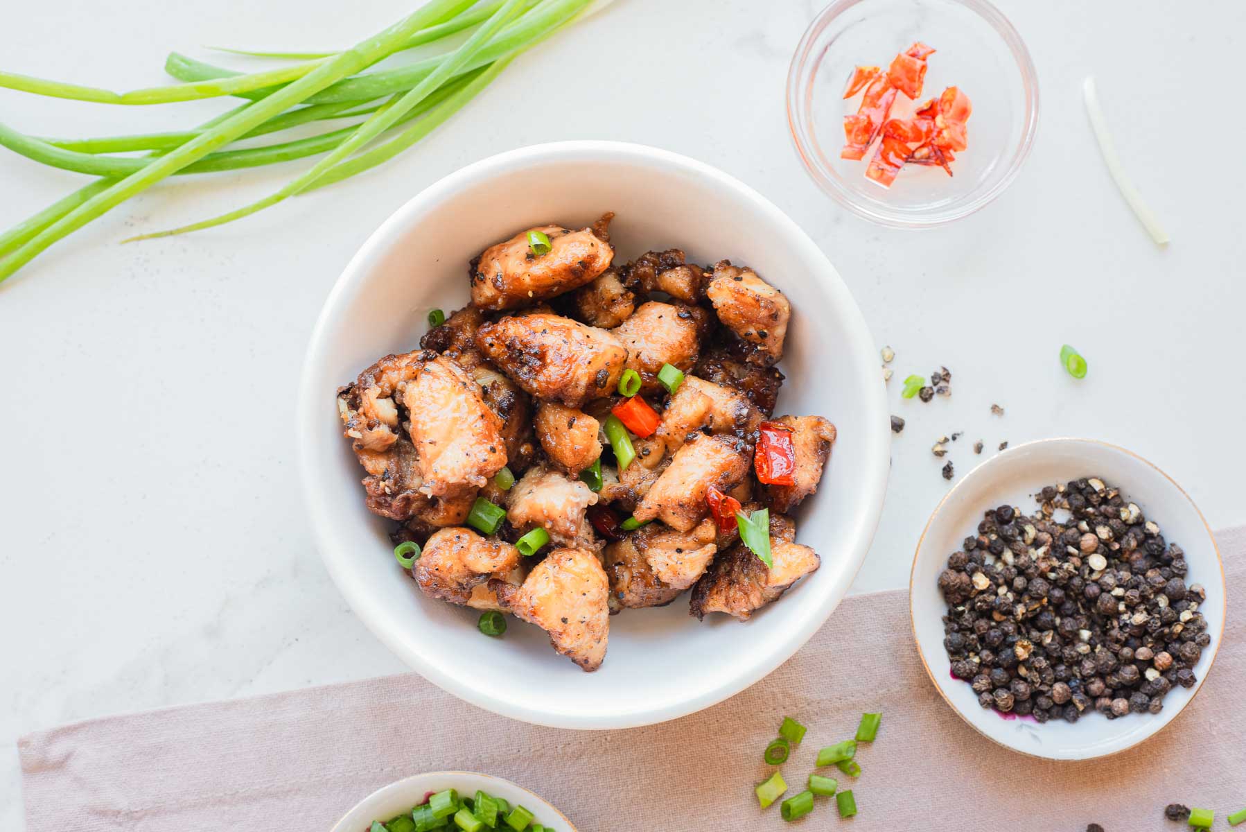 A bowl of Air Fryer Salt and Pepper Chicken garnished with green onions, surrounded by pepper, green onions, and red chili.