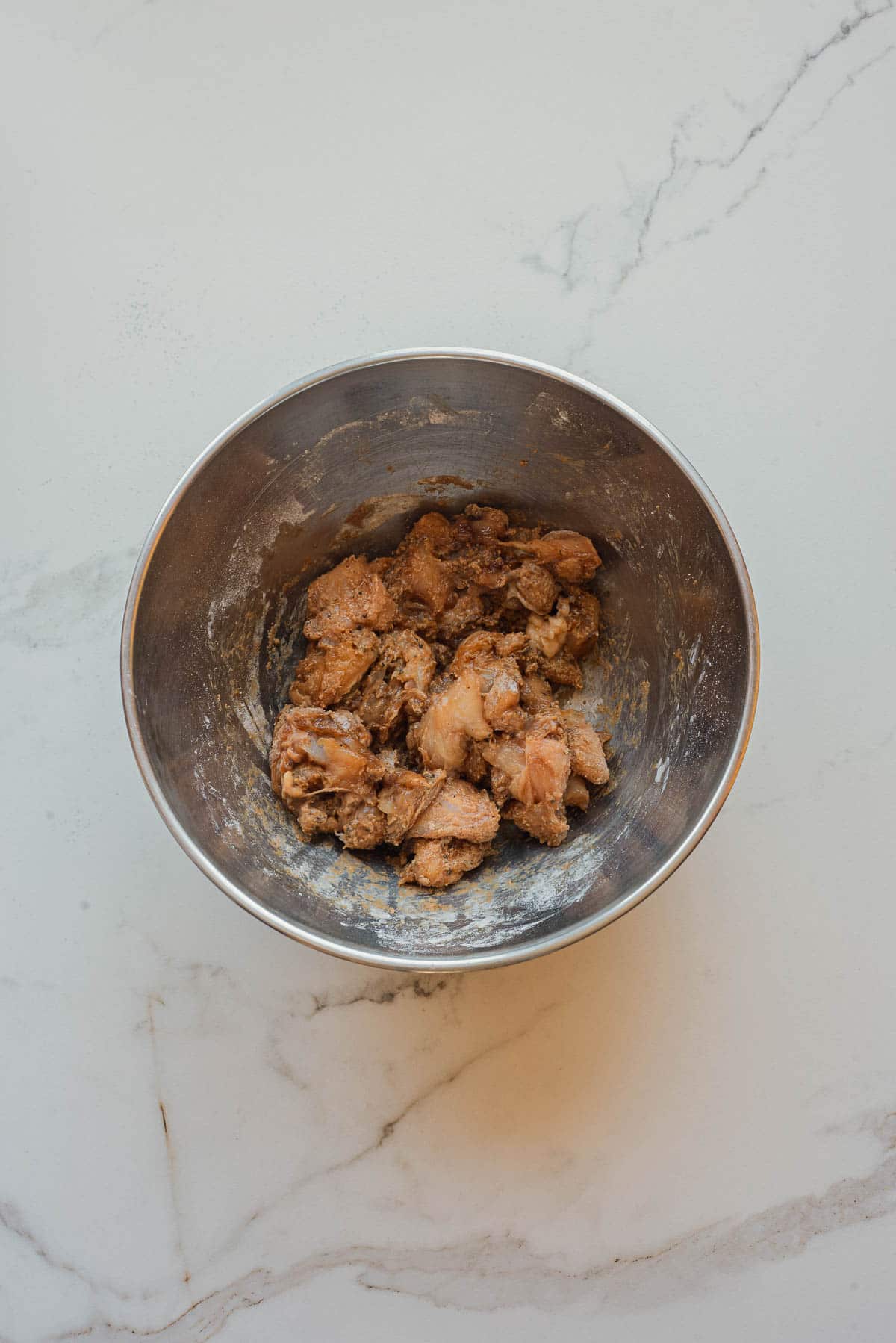 A metal bowl with pieces of marinated raw chicken, perfect for a quick Salt and Pepper Chicken recipe, sits on a white marble surface.