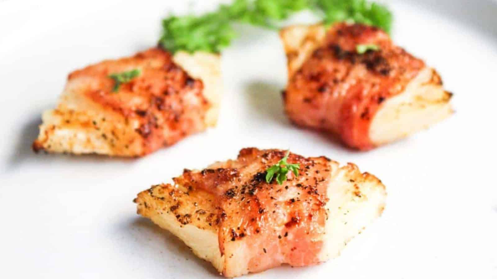 Three pieces of bacon-wrapped fish garnished with herbs on a white plate.