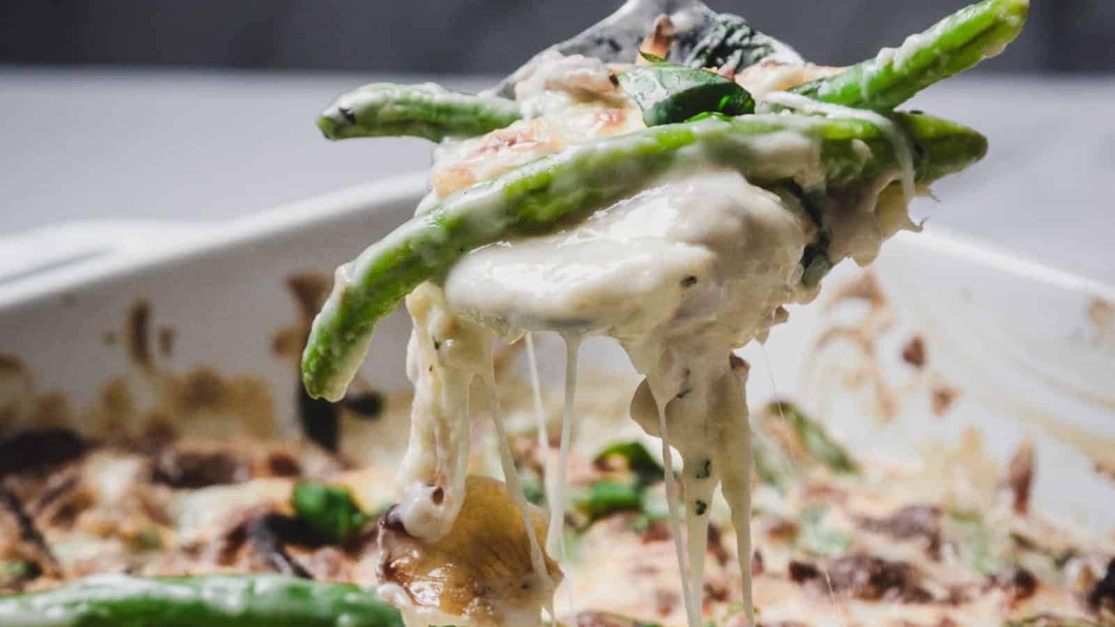 A cheesy green bean casserole being served from a white baking dish with a spoon.
