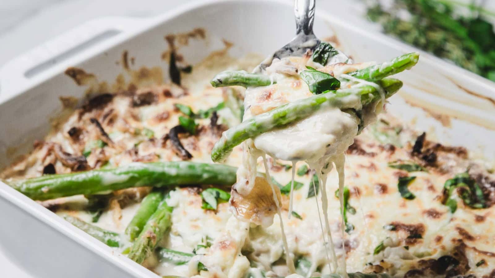A spoon lifts cheesy green bean casserole from a white baking dish, topped with melted cheese and herbs.