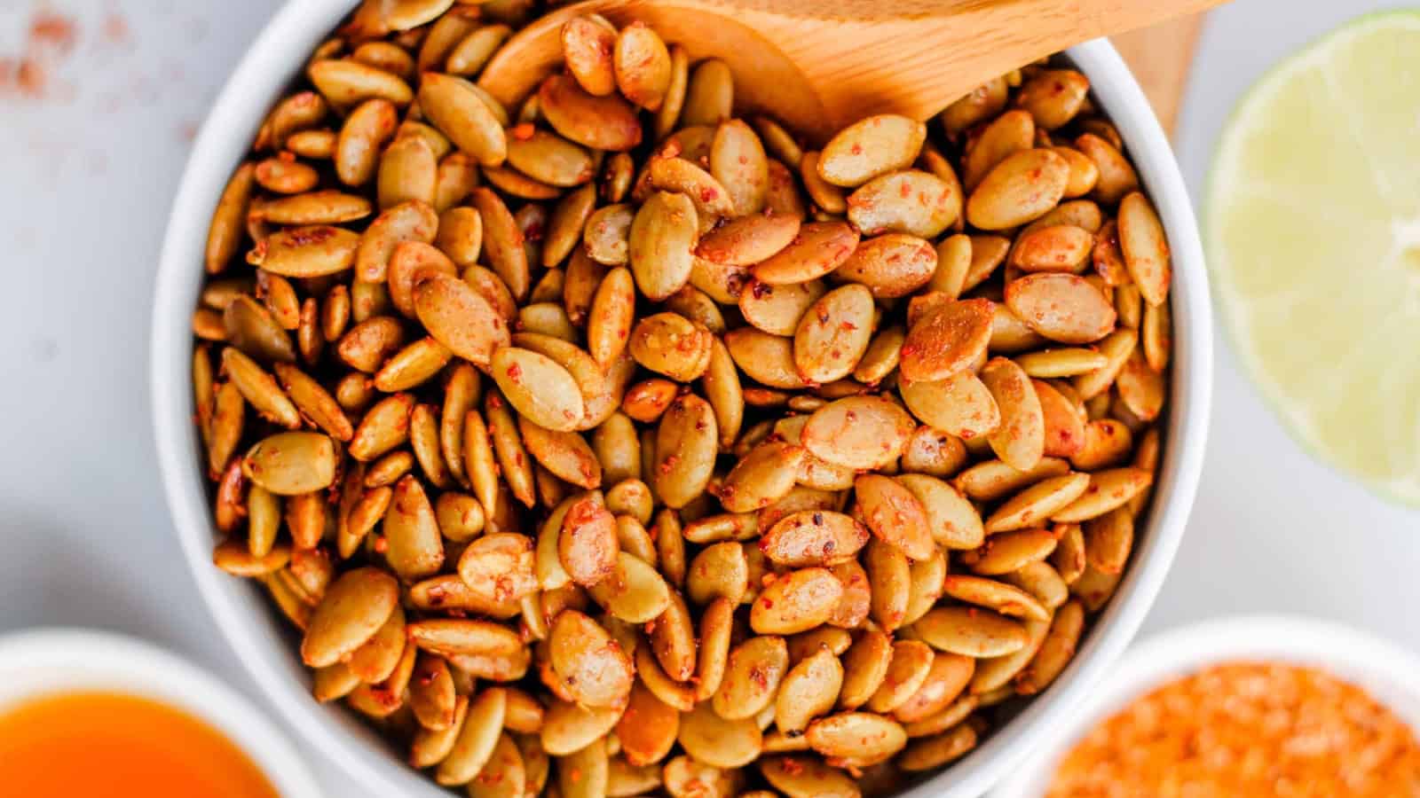A bowl of seasoned pumpkin seeds with a wooden spoon, next to a lime slice.