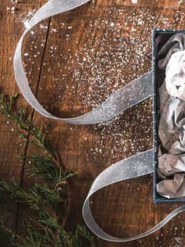 A gift box with three powdered sugar cookies on crumpled paper, ribbon, and holiday decor on a wooden table.