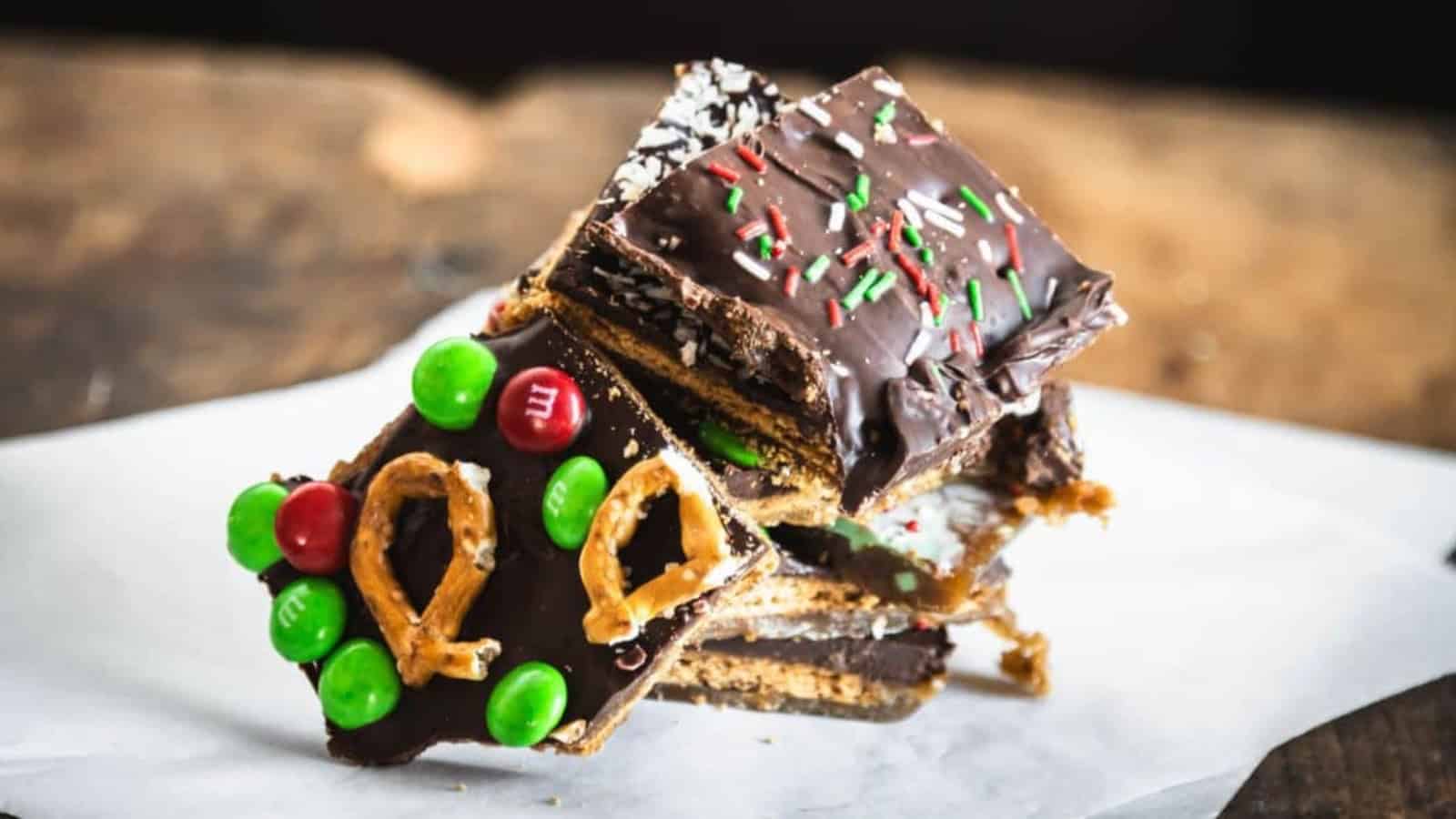 Stack of chocolate bark topped with pretzels, green and red candies, and holiday sprinkles on parchment paper.