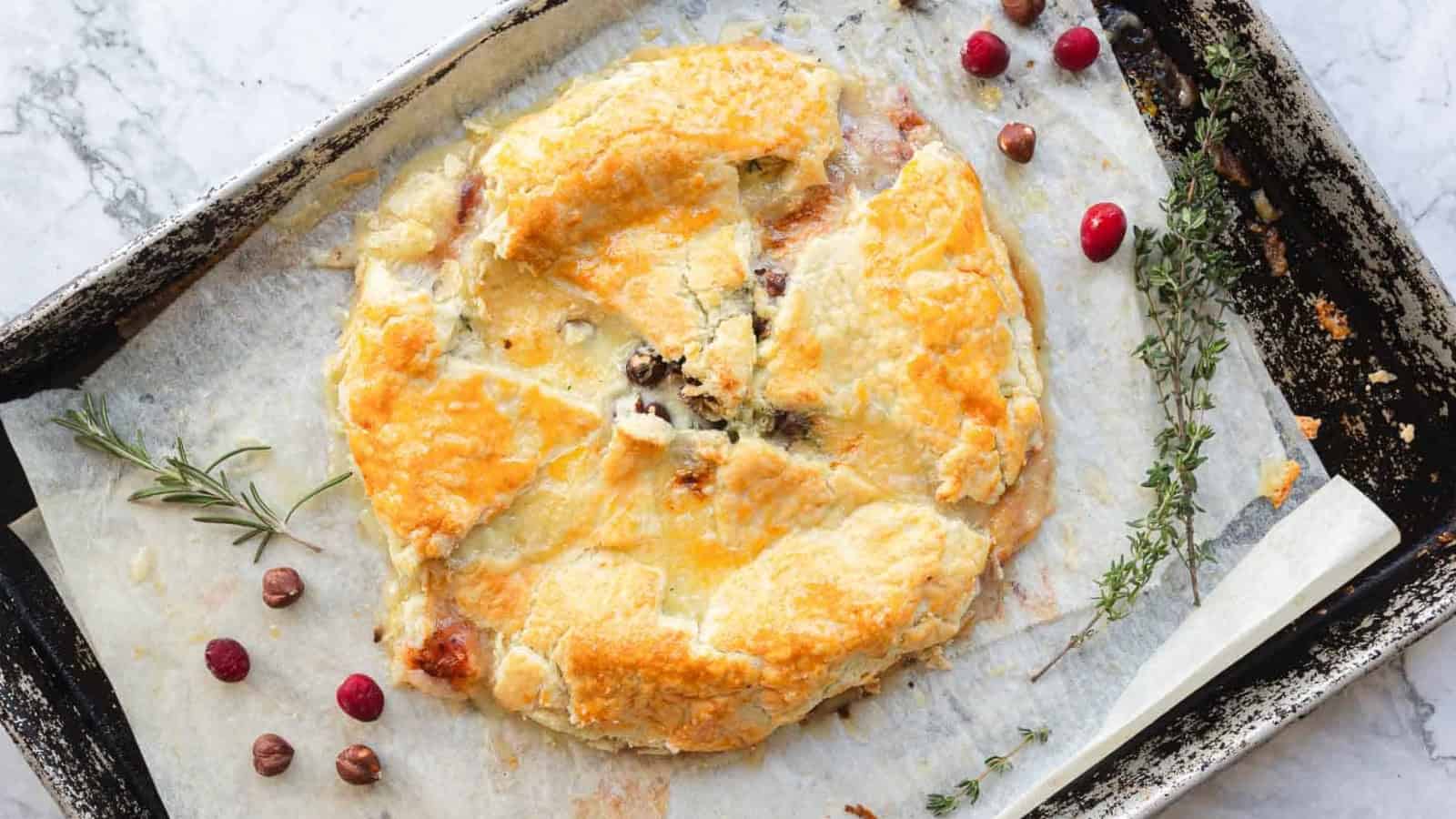 Golden baked pastry on parchment with herbs, cranberries, and nuts, on a rustic baking sheet.