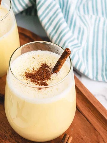 Two glasses of eggnog topped with cinnamon sticks on a wooden tray, with striped cloth in the background.