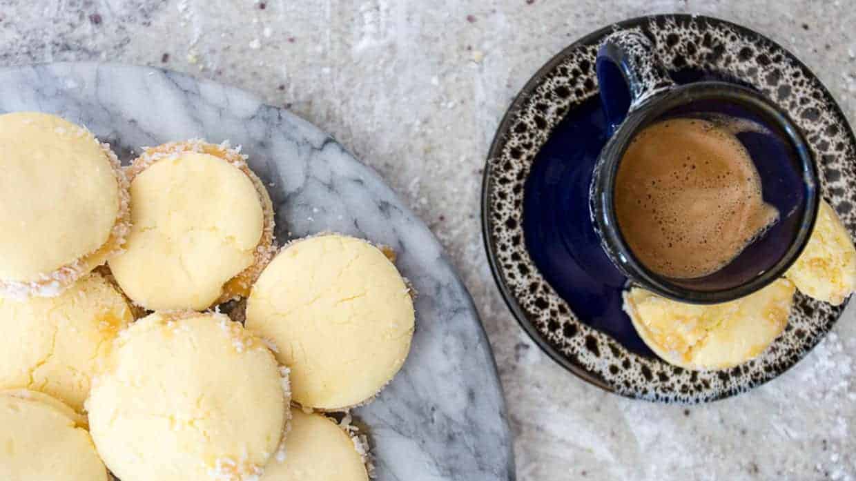 A marble plate of yellow cookies next to a cup of coffee with a cookie on the saucer.