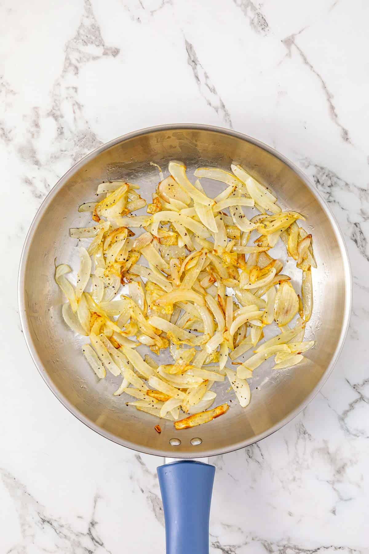 Sliced onions sautéing in a stainless steel pan with a blue handle on a marble countertop—perfect for adding rich flavor to your BBQ Chicken Sliders.