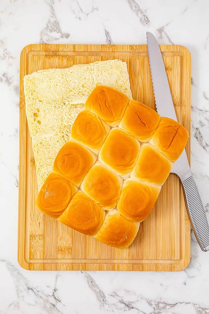 A bread knife and a sliced sheet of dinner rolls on a wooden cutting board, ready to be filled for delicious BBQ Chicken Sliders, on a marble surface.